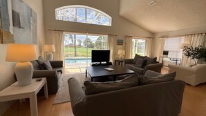 A spacious living room with a large TV, comfortable seating, and a view of a swimming pool. - 8th Fairway Pool Villa, Southern Dunes (Haines City)
