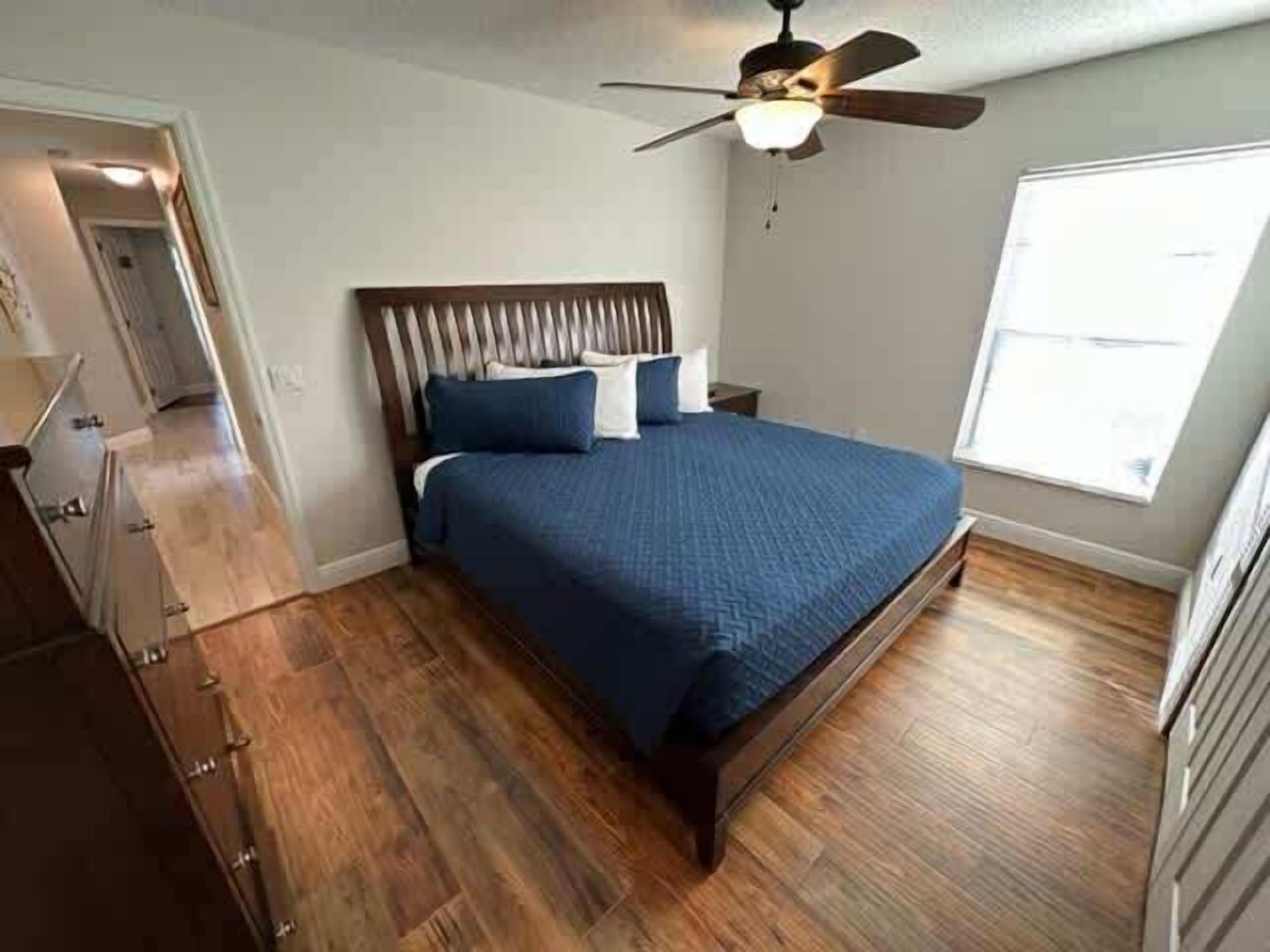A bedroom with a wooden floor, a bed with blue bedding, a ceiling fan, and a window.