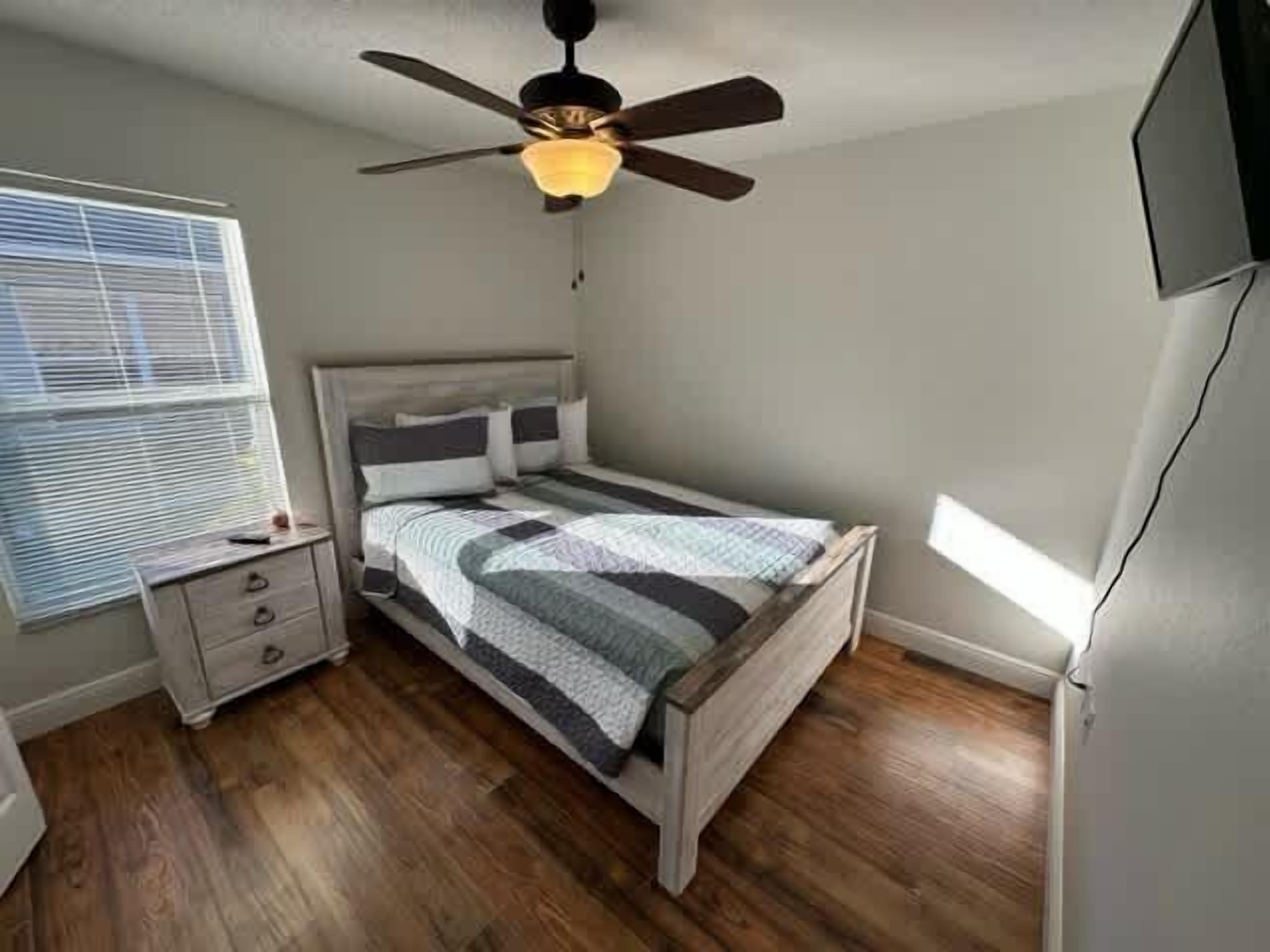 A bedroom with a bed, a nightstand, a dresser, a ceiling fan, and a window with blinds.