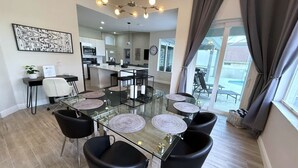 A modern dining area with a glass table surrounded by black chairs, a chandelier, and a view of the kitchen and outdoor patio. - Elite Forest Hills Villa - Southern Dunes - Golf Course Views (Haines City)