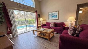 A spacious living room with a large glass door leading to a pool, a wooden coffee table, and a red sofa. - Private Pool Villa, Many Updates! Southern Dunes (Haines City)