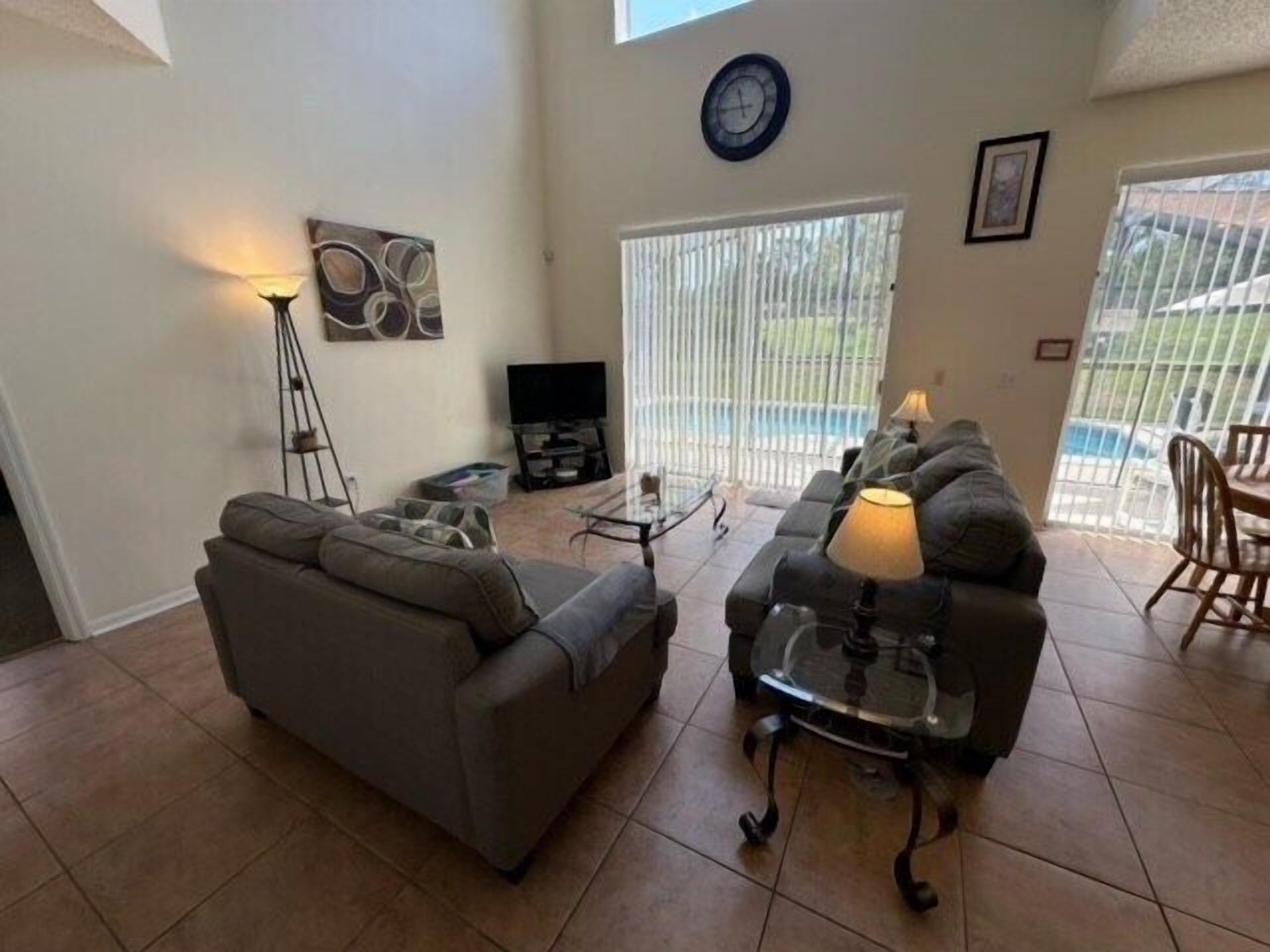 A living room with a gray sofa, a glass coffee table, and a TV. There is a large window with blinds, a clock on the wall, and a painting.