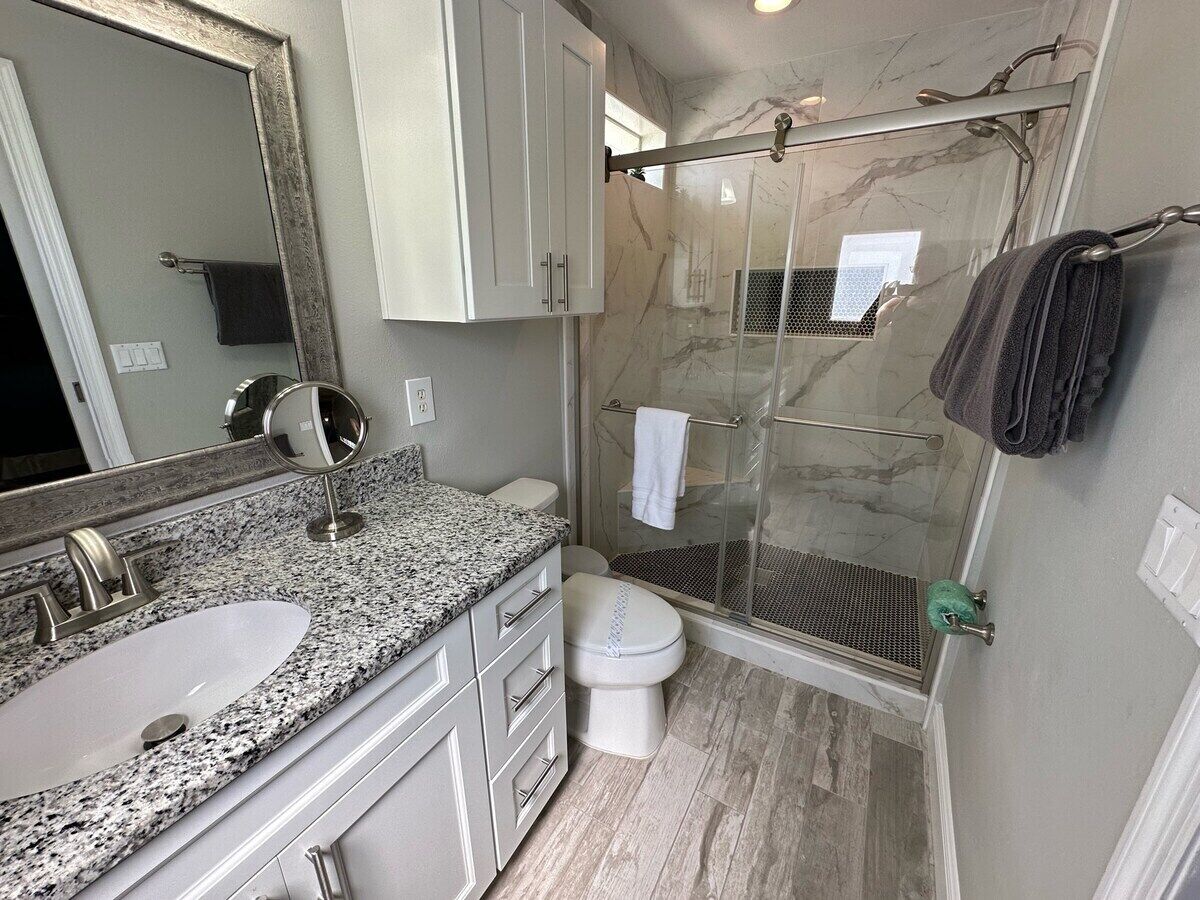 A bathroom with a granite countertop sink, a toilet, a glass shower enclosure, and a large mirror.