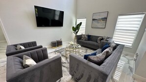 A modern living room with grey sofas, a flat-screen TV, a coffee table, and a large window with blinds. - Beautifully Remodeled Home - Southen Dunes Golf Course Views (Haines City)