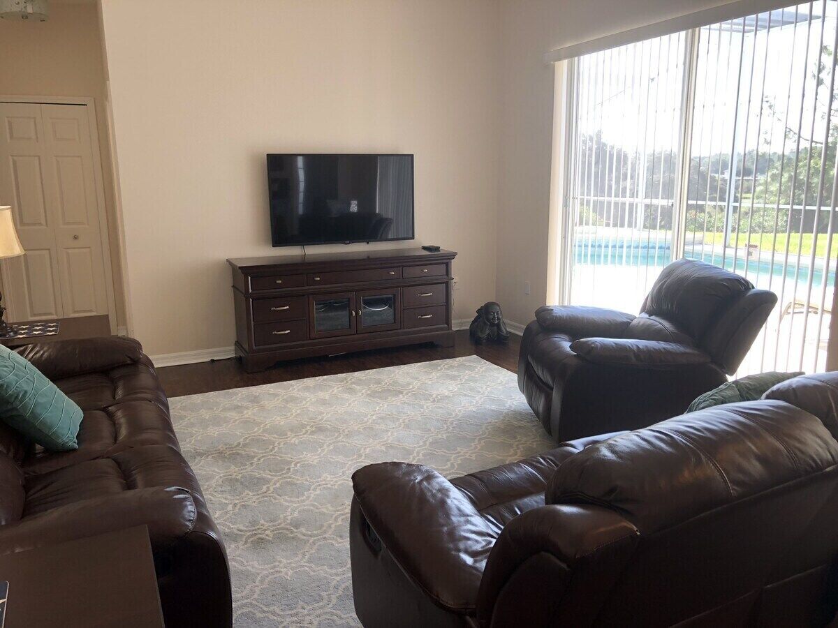 Un salón con un sofá de cuero marrón, un televisor sobre un mueble de madera y un ventanal con vistas a una piscina.