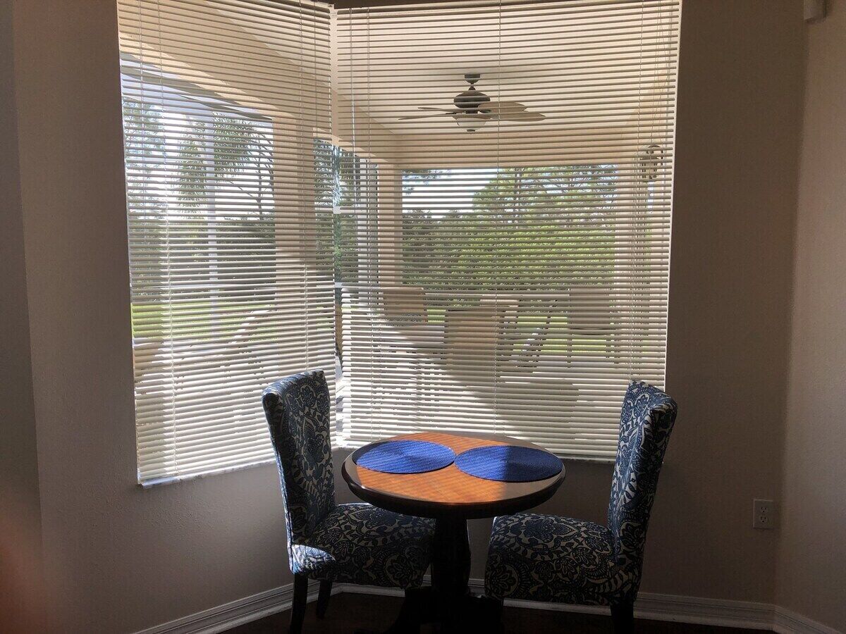 A room with a round table and two patterned chairs, large windows with blinds, and a ceiling fan.