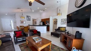 A spacious living room with a sofa, coffee table, and a TV. There is a kitchen area in the background. - South Facing Golf Course Views, Southern Dunes Villa (Haines City)