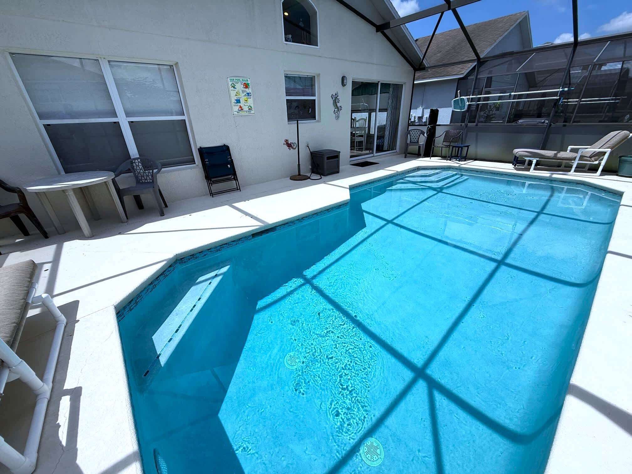 Une piscine rectangulaire à l’eau d’un bleu limpide, entourée de murs blancs et dotée d’un patio couvert avec des chaises et des tables.