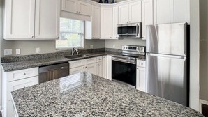Una cocina con armarios blancos, encimeras de granito, electrodomésticos de acero inoxidable y un ventanal con persianas.