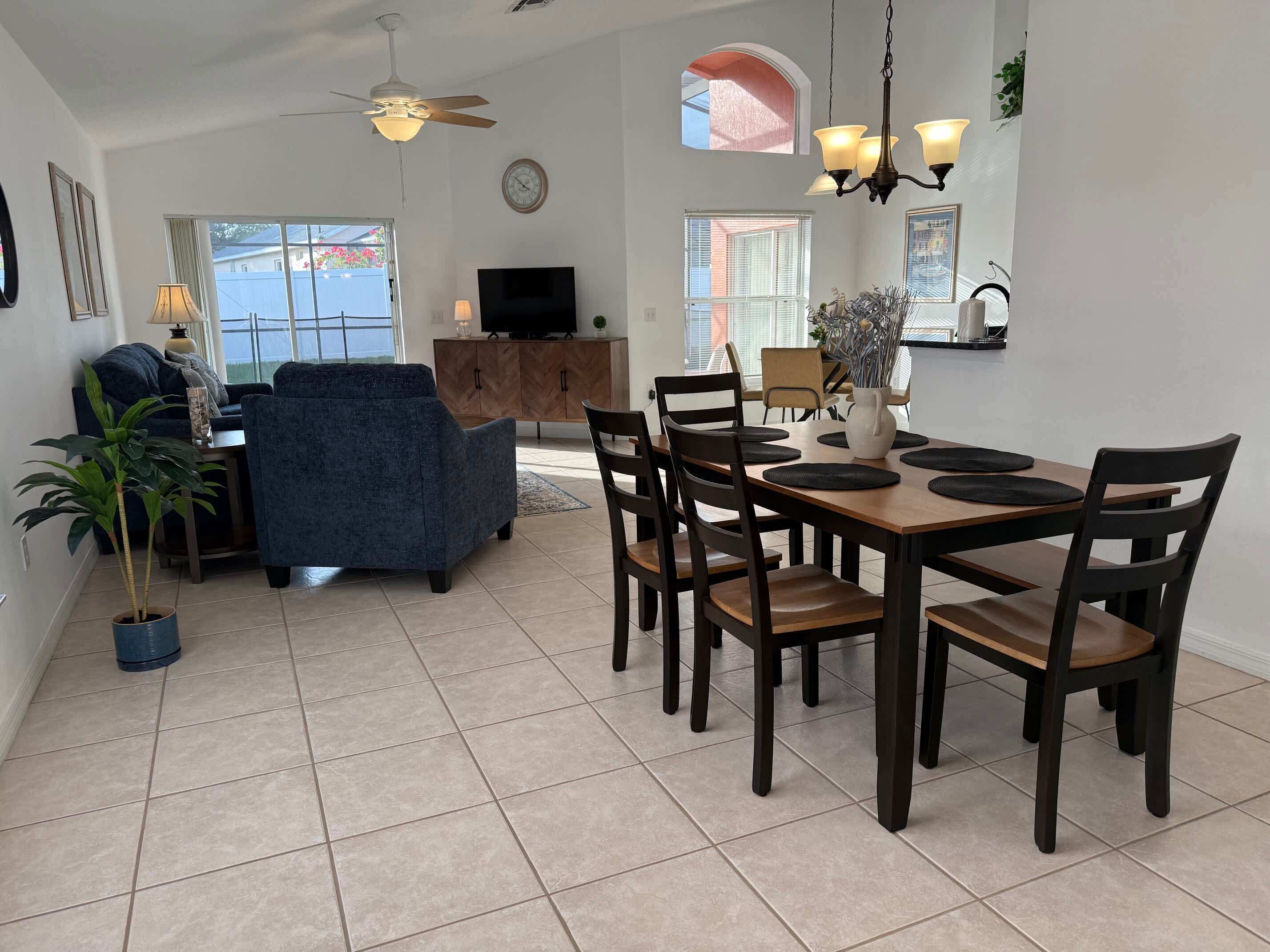 A spacious living room with a dark blue sofa, a dining area with a wooden table and chairs, a ceiling fan, and a TV in the background.