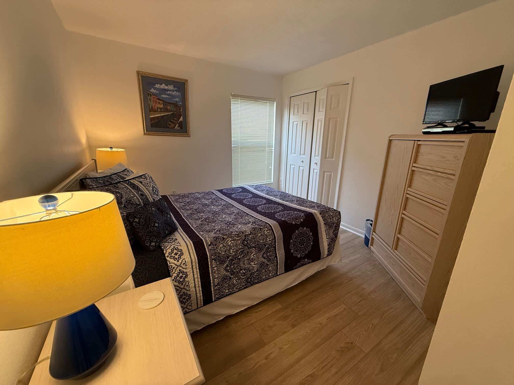 Un dormitorio con cama, mesita de noche con lámpara, cómoda, televisor sobre un mueble bajo y ventana con persianas.