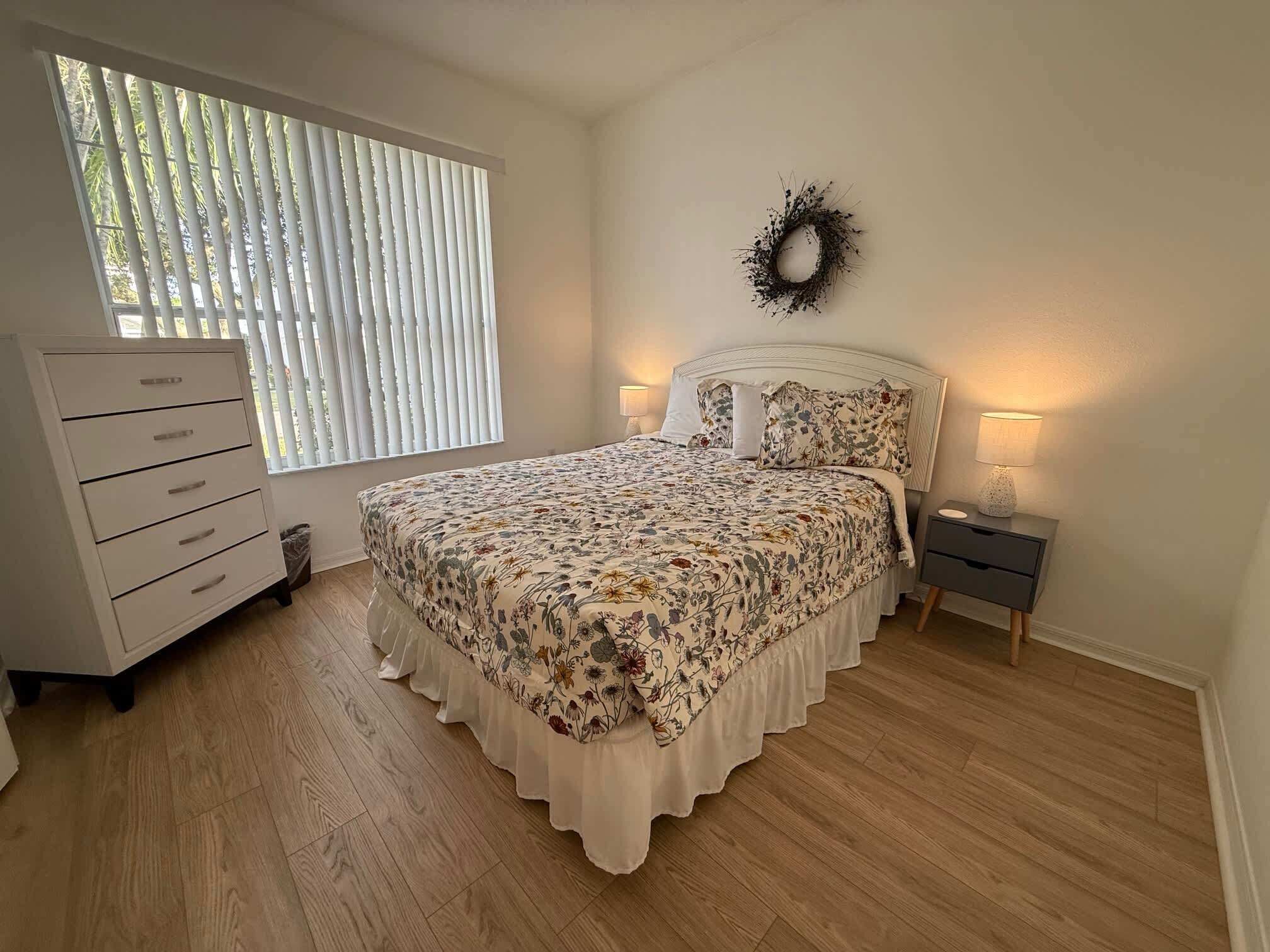 A bedroom with a bed, a dresser, a nightstand, a wall-mounted wreath, and a window with blinds.