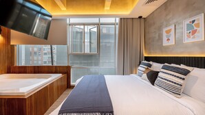 Ein modernes Hotelzimmer mit einem großen Bett, einer integrierten Badewanne, einem großen Fenster mit Blick auf Gebäude und abstrakter Kunst an der Wand.