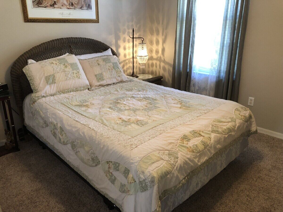 Un lit bien fait, recouvert d’une courtepointe à motifs floraux, une table de chevet avec une lampe et un tableau encadré accroché au mur.