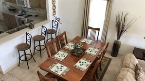 Une salle à manger avec une table en bois dressée pour six personnes, entourée de chaises. On aperçoit une cuisine au fond.