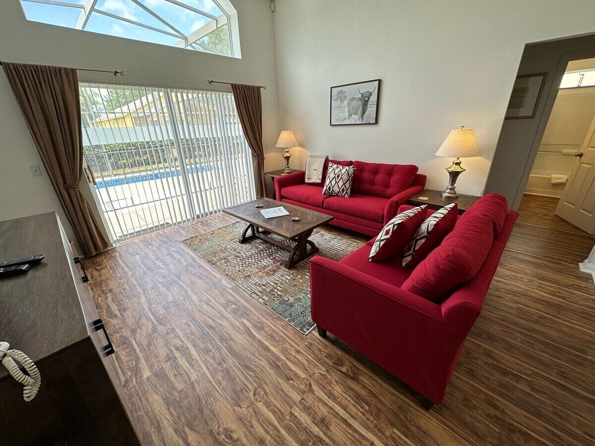 Ein Wohnzimmer mit einem roten Sofa, einem Couchtisch und zwei Lampen. Ein Fenster mit Vorhängen bietet Blick auf einen Zaun.