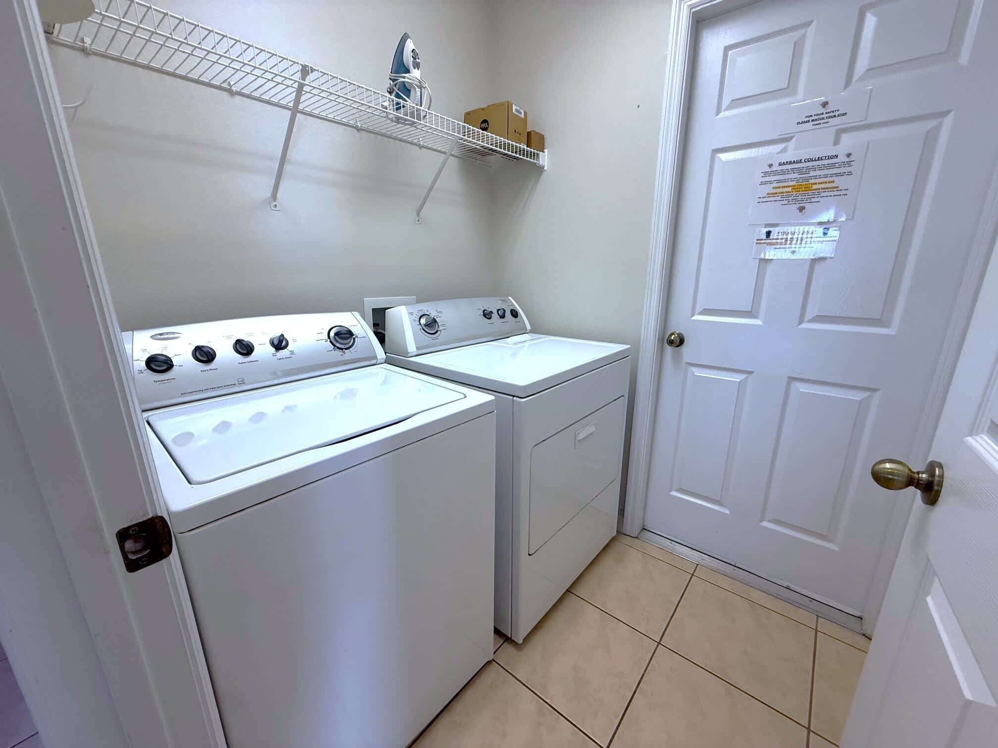 A laundry room with two white washers and dryers, a metal shelf above, and a door with a garbage collection sign.