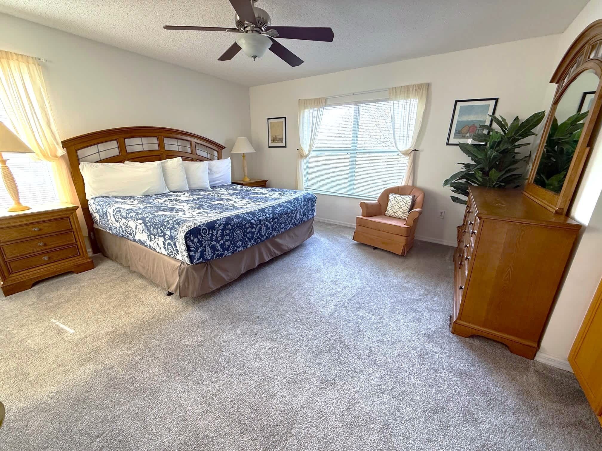 Une chambre à coucher comprenant un lit, une chaise, une commode, un ventilateur de plafond et une fenêtre avec des rideaux.