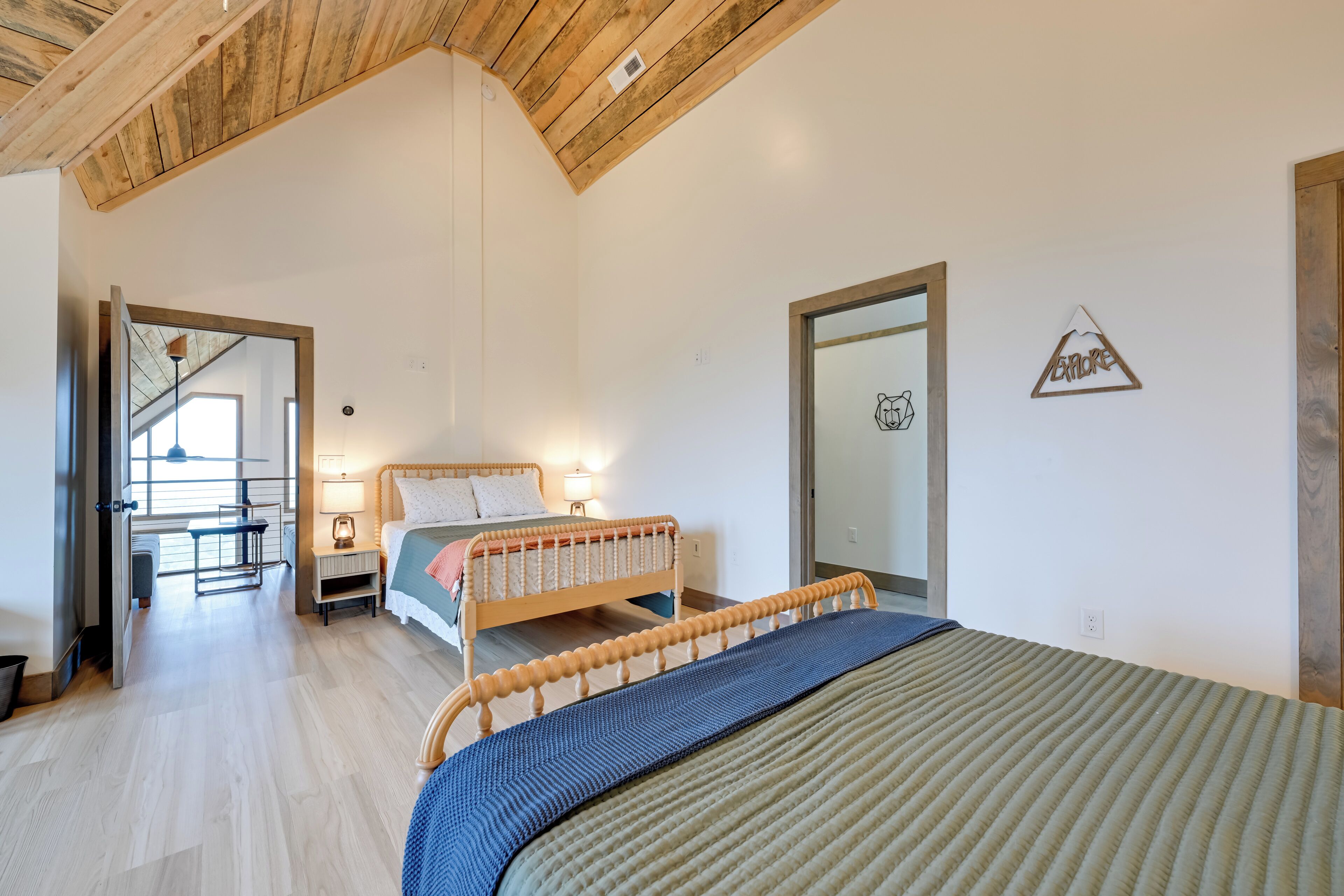 Ein Schlafzimmer mit Holzbalkendecke, einem Bett mit blau-beiger Bettdecke, einem Nachttisch mit Lampe und einem Fenster mit Blick ins Freie.