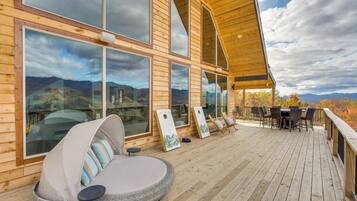 Une terrasse en bois dotée d’une grande fenêtre donnant sur un paysage montagneux. On y trouve un espace salon circulaire avec des coussins, ainsi qu’une table entourée de chaises.