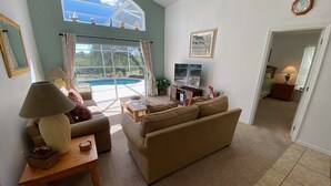 A living room with a sofa, coffee table, lamp, and a view of a swimming pool through a large window. - Golf Course Views, South Facing Pool Villa (Haines City)