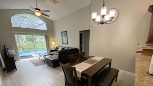 A spacious dining area with a wooden table and chairs, a ceiling fan, and a view of the outdoors through a large window. - Southern Dunes Villa, Upgraded, Golf Views (Haines City)