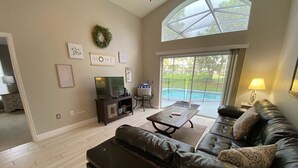 A spacious living room with a leather sofa, a coffee table, and a TV. There is a large window with a view of a swimming pool. - Southern Dunes Villa, Upgraded, Golf Views (Haines City)