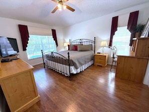 Quarto com cama, mesas de cabeceira, escrivaninha, ventoinha no teto e janelas com cortinas.