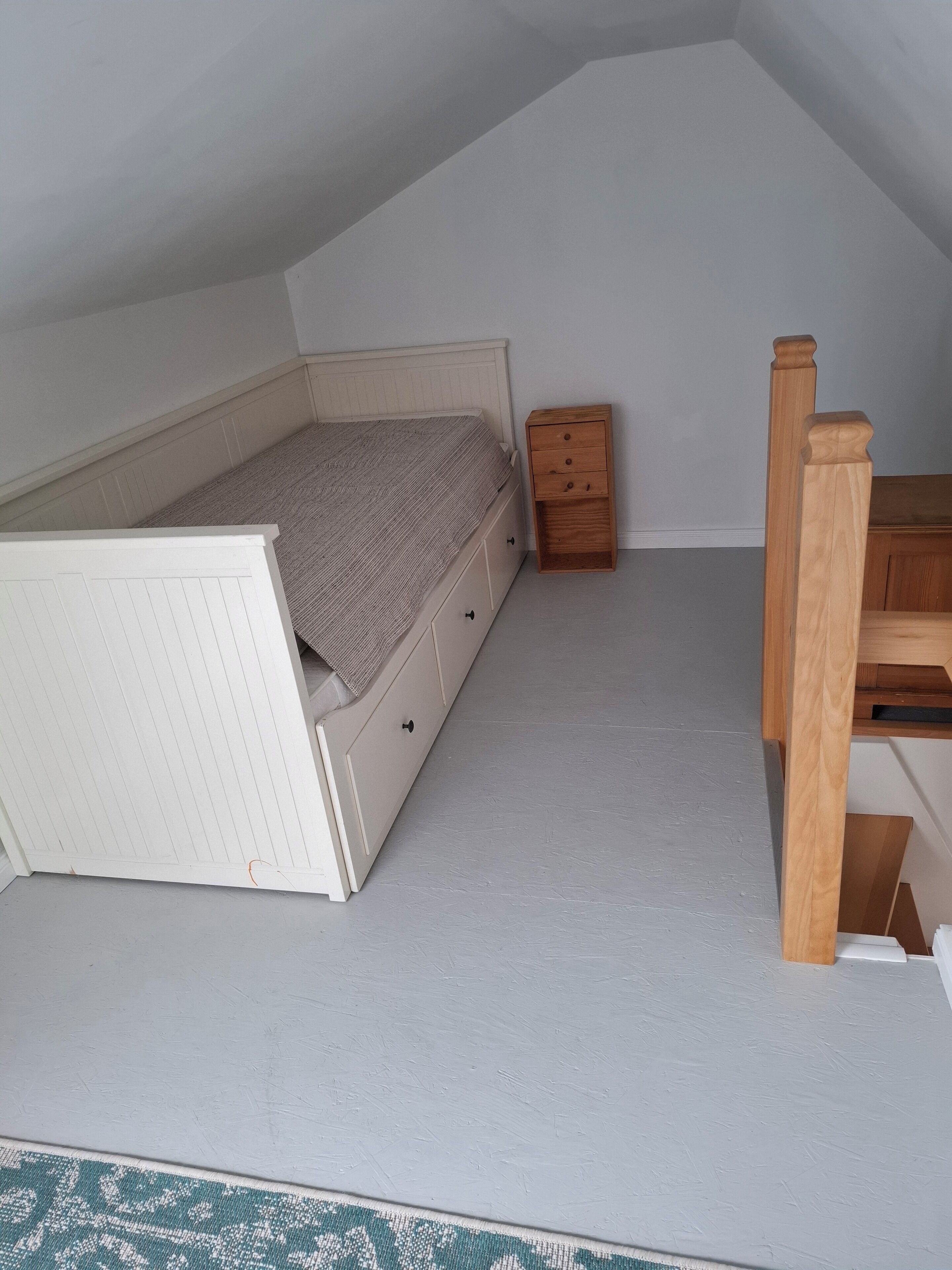 A small, sloped ceiling room with a bed, a wooden nightstand, and a desk.