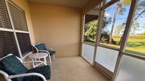 A balcony with two chairs and a small table, a beige wall, and a view of a green landscape. - Southern Dunes Condo, Golf Course Views (Haines City)