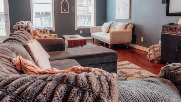 Una sala de estar acogedora con un amplio sofá modular, un sillón blanco pequeño y una mesa de centro. Hay una chimenea y una ventana con persianas.