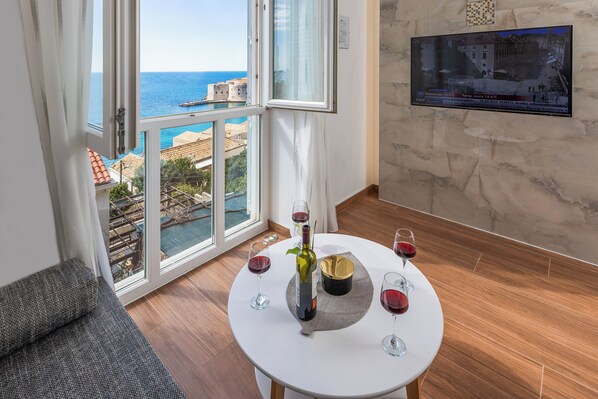 Une pièce dotée d’une grande fenêtre donnant sur la côte, une table ronde avec des verres à vin et une bouteille, et un panneau d’information fixé au mur.