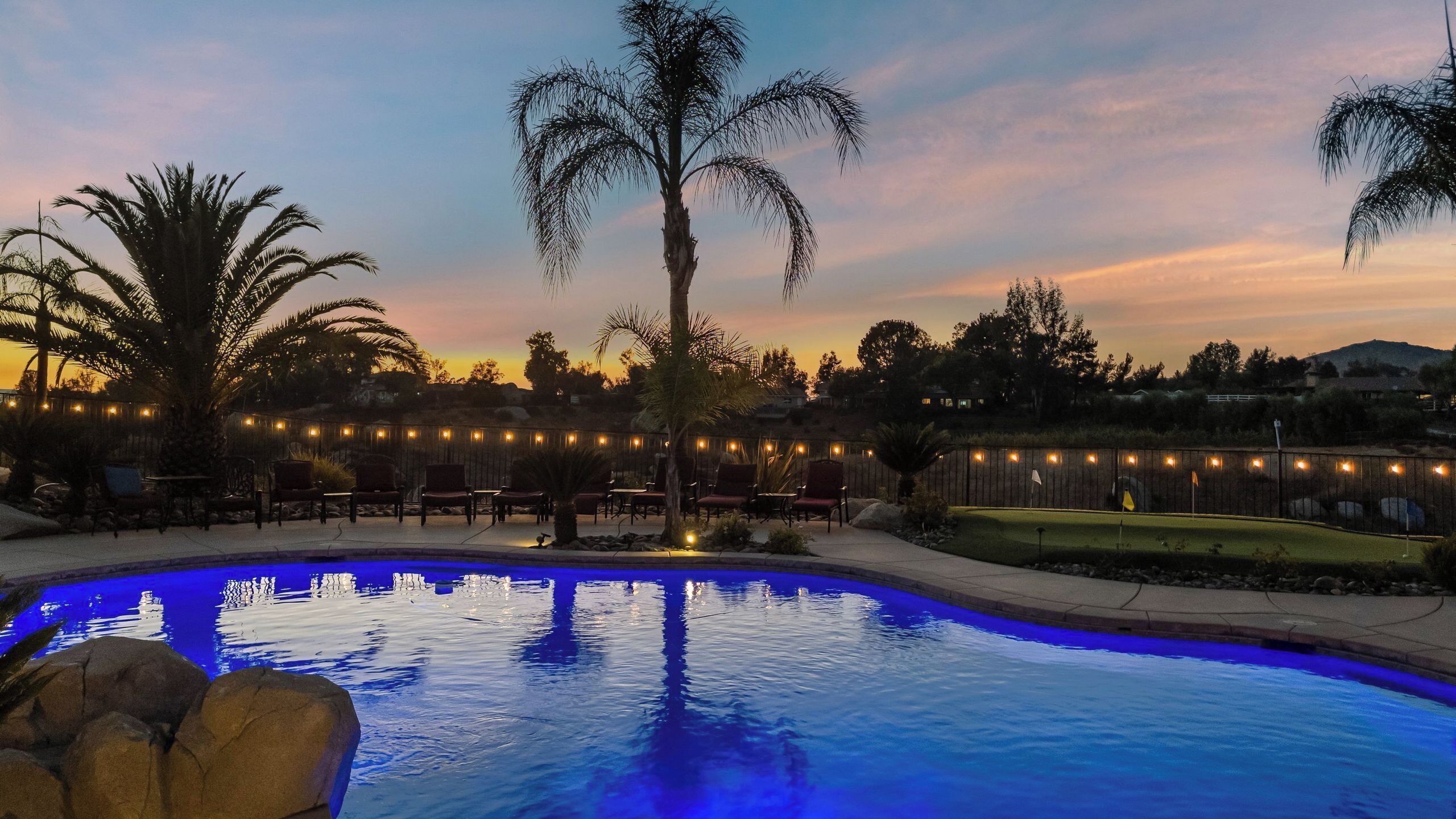 Une piscine dont le bord est illuminé de lumière bleue, entourée de palmiers et délimitée par une clôture en bois ornée de guirlandes lumineuses.