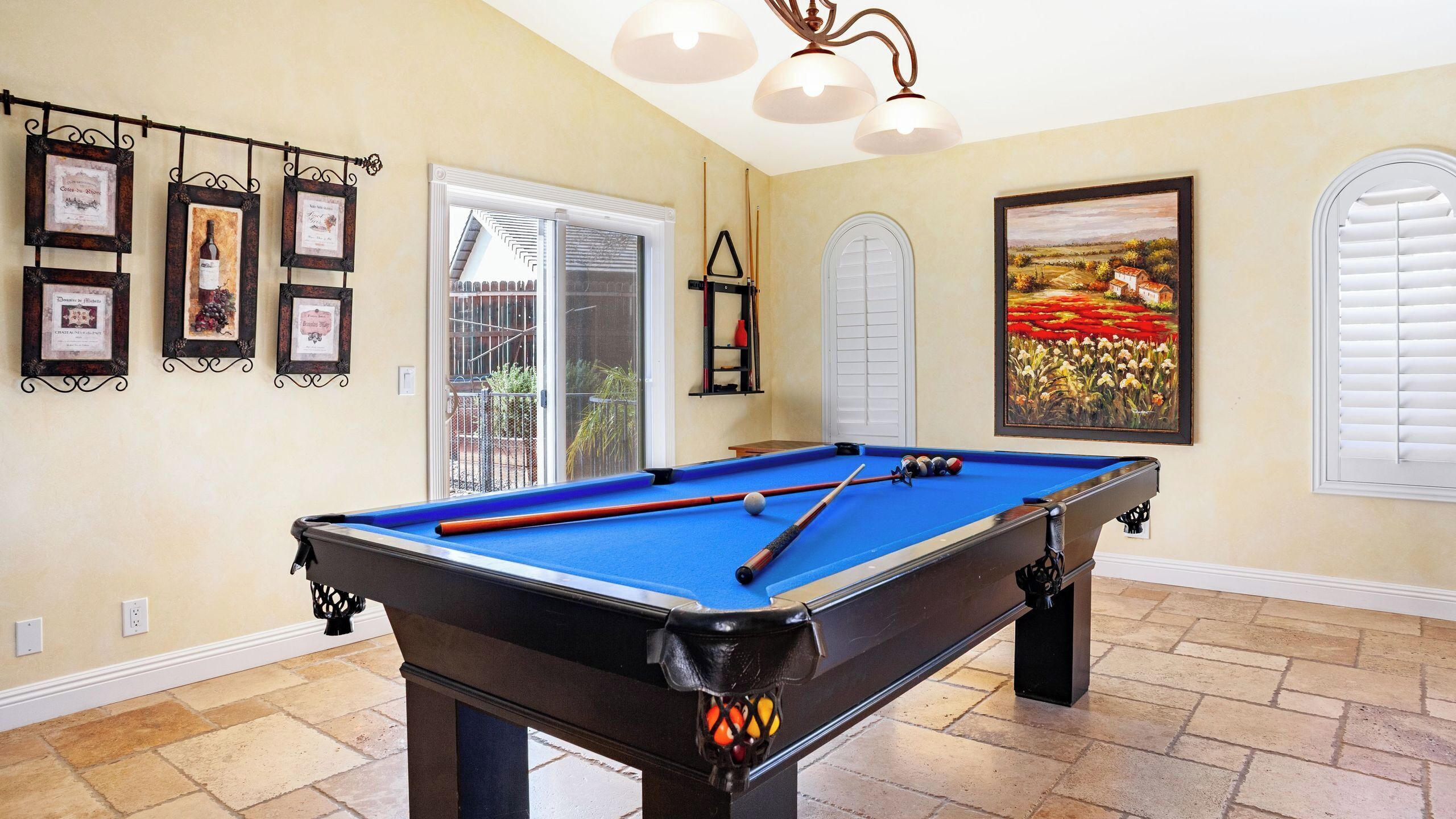 A pool table with a blue felt surface, a billiards cue, and a red cue ball. There are framed certificates on the wall, a painting of a field with flowers, and a window with white blinds.