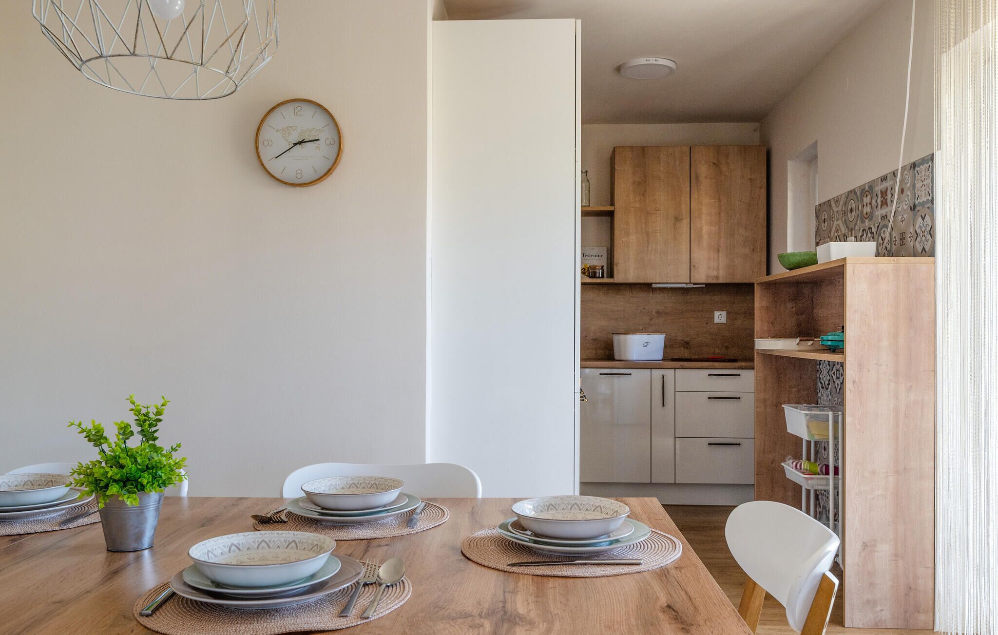 Un coin repas avec une table en bois dressée pour quatre personnes, une plante en pot et une horloge murale.