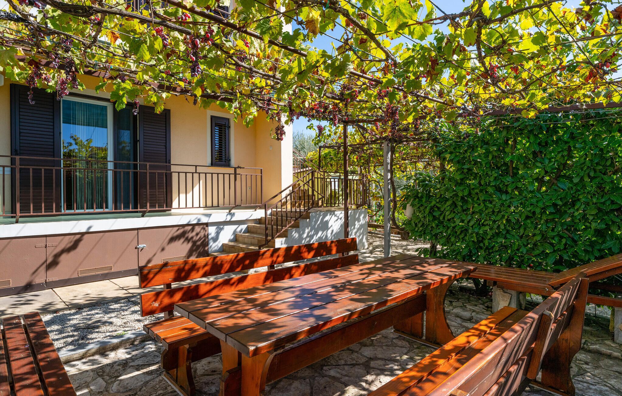 Eine Terrasse mit Holzbänken und einem Tisch, umgeben von einem gelben Gebäude mit Fensterläden und einer Treppe zu einem Balkon.
