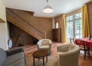 Amplia sala de estar con una escalera de madera alta, un comedor con mantel rojo y vistas al exterior a través de amplias ventanas.