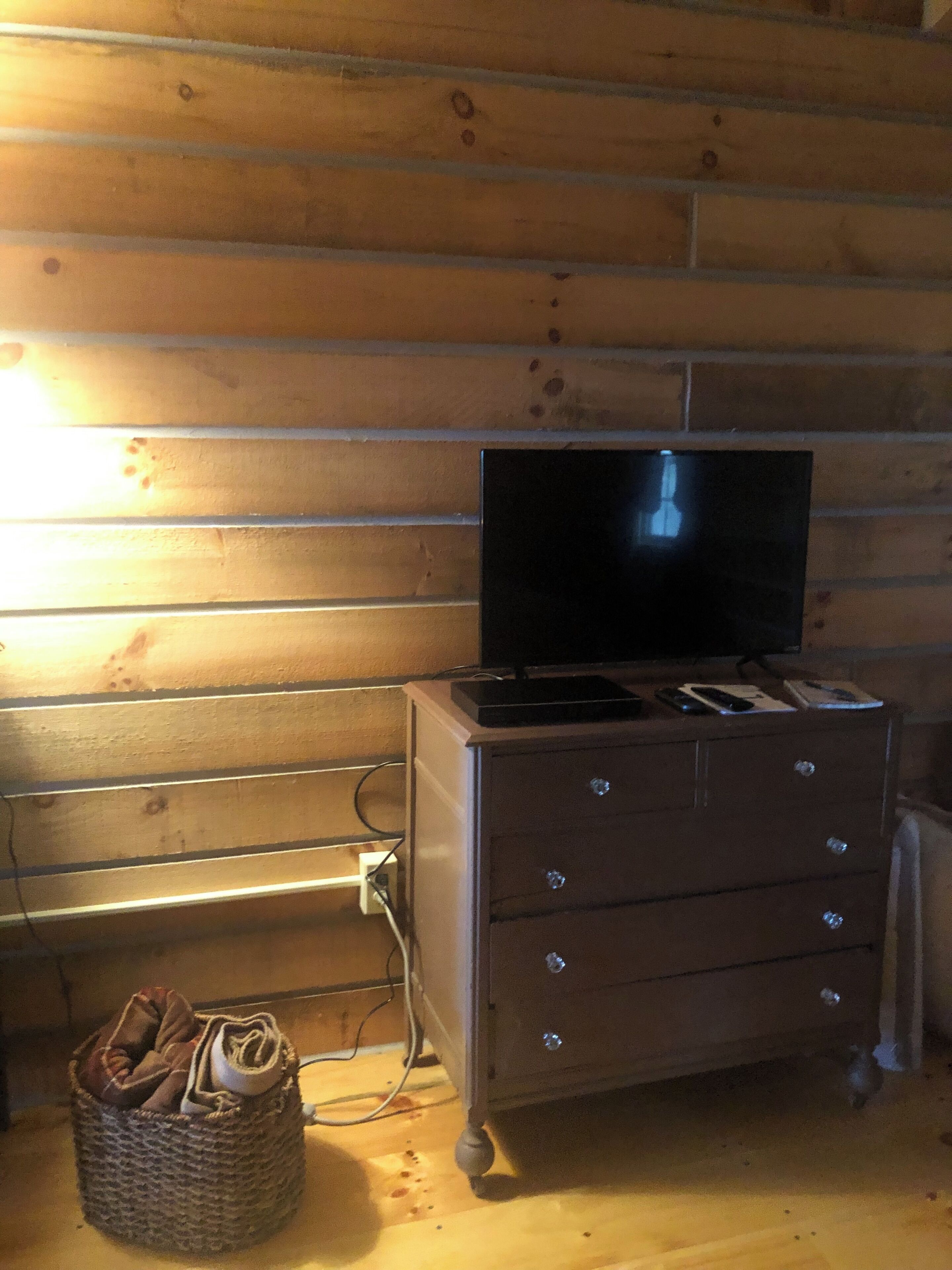 Een houten blokhut van binnen, met een televisie op een dressoir, een mand met kleding en een stopcontact.