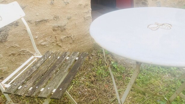 Een rustieke buitenruimte met een ronde tafel en een houten bankje, een rode deur en een ruwe stenen muur.