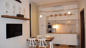 A compact kitchen with white cabinets, a microwave, and a sink. There are two white chairs at a small table, and a flat-screen TV mounted on the wall.
