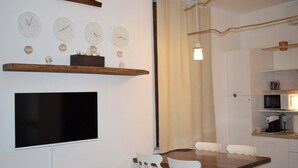 A compact kitchen with a dining area, a flat-screen TV, and wall clocks.