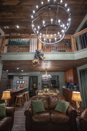 A spacious living area with a high ceiling, a wooden ceiling, a chandelier, a leather sofa, and a dining area with stools and a table. - Trailhead Lodge • Sleeps 8 • 2 Kings + Bunks • Game Room • Steps to Trail (Gatlinburg)