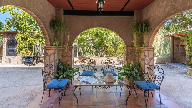 Un patio exterior con una mesa de vidrio, cuatro sillas de metal con cojines azules y plantas en macetas.