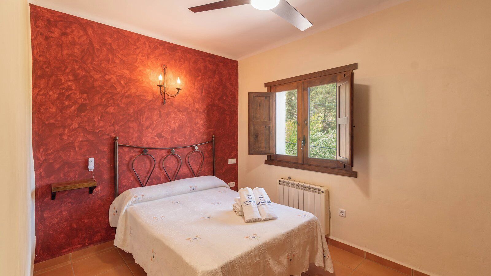 A bedroom with a bed, a wall-mounted light, a window with wooden shutters, and a radiator.