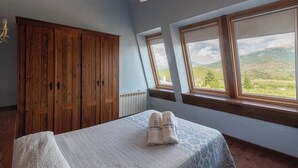 A bedroom with a large wooden wardrobe, a bed with a patterned bedspread, and a view of a green landscape through a window.
