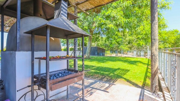 Un barbecue extérieur au charbon de bois, une table en métal avec des légumes et une clôture métallique entourant une zone herbeuse.