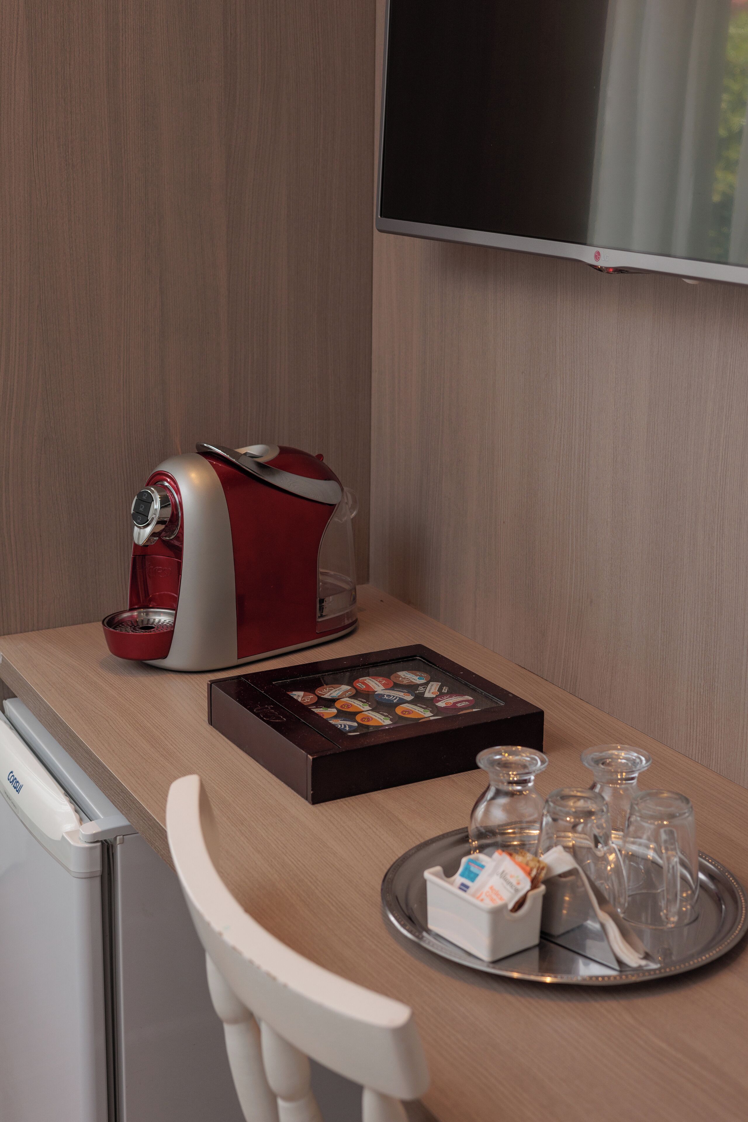 Una habitación de hotel moderna con un escritorio de madera, una cafetera roja y blanca, una bandeja con vasos y aderezos, y un televisor de pantalla plana.