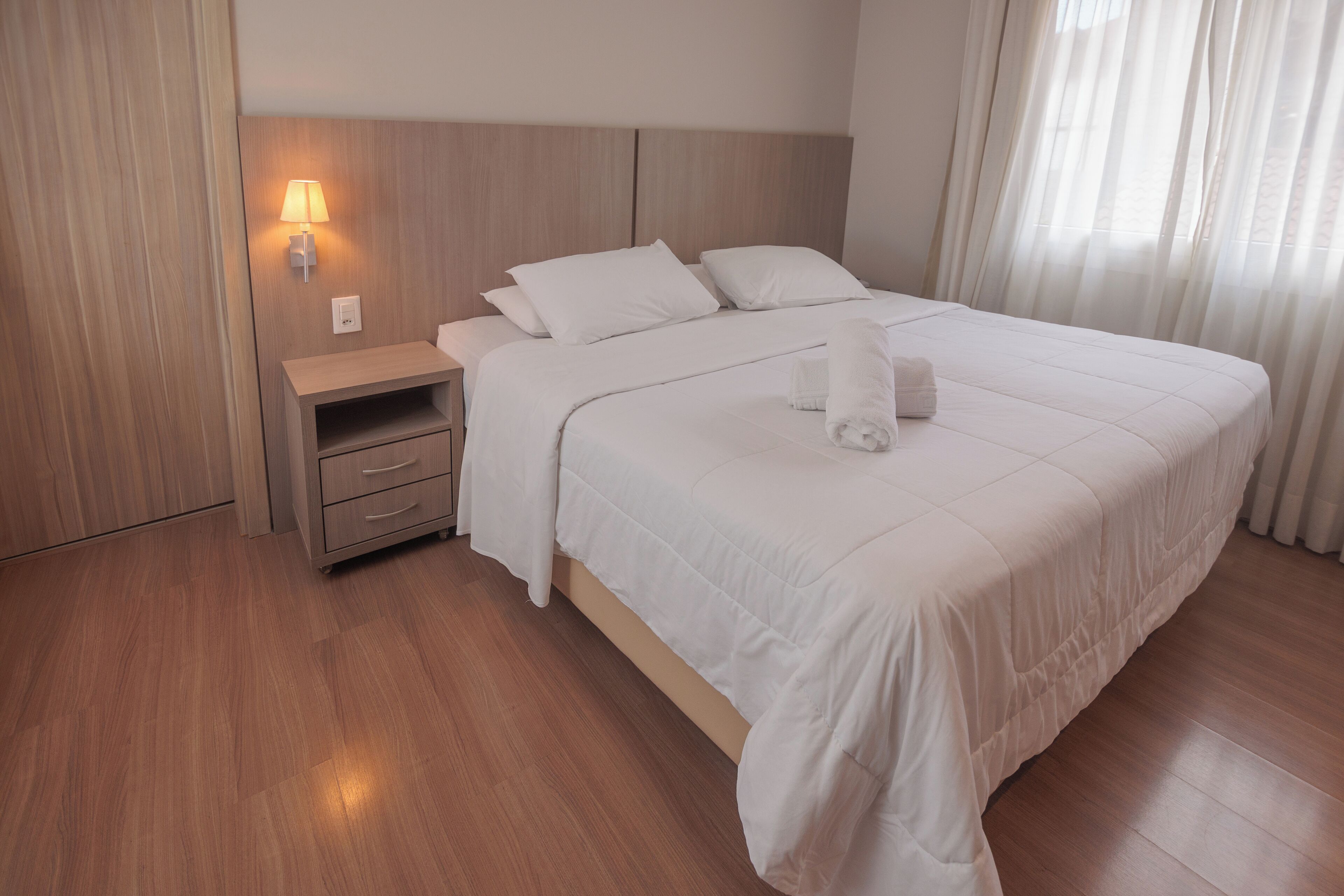 Una cama bien hecha con ropa blanca, una lámpara de noche y una mesita de noche de madera con cajones.