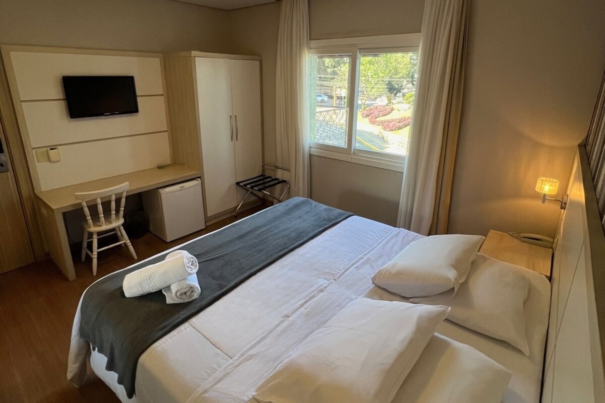 Une chambre d’hôtel avec un lit, un bureau, une chaise, une télévision et une fenêtre avec des rideaux.