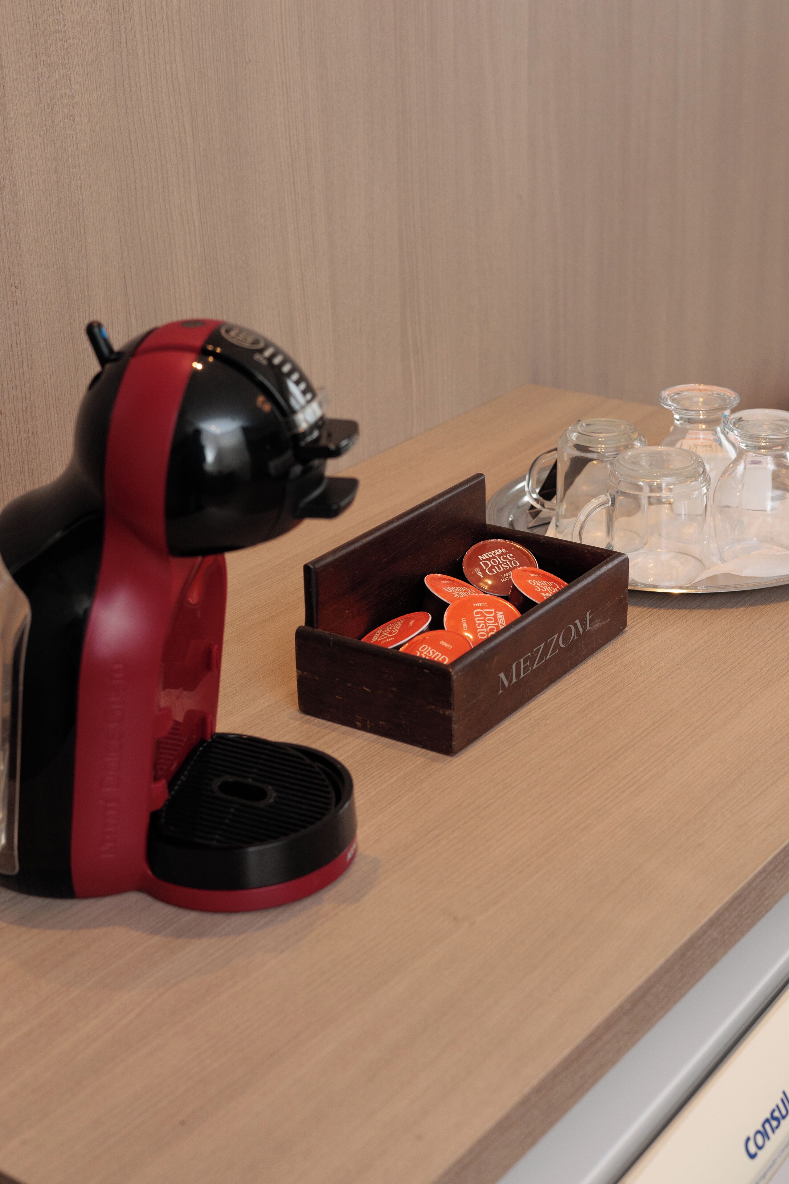A red and black coffee maker, a box of coffee pods, and a tray with glass mugs on a wooden table.